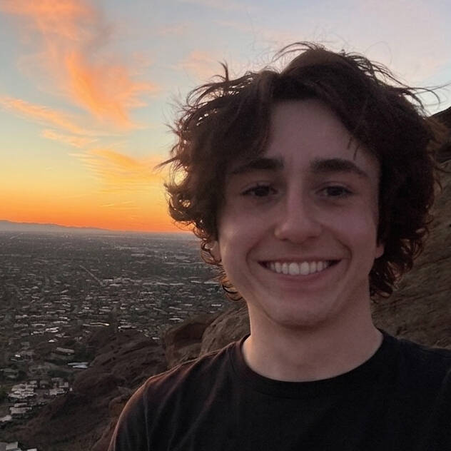 Smiling young man with curly brown hair in the foreground with a sunset horizon in the background